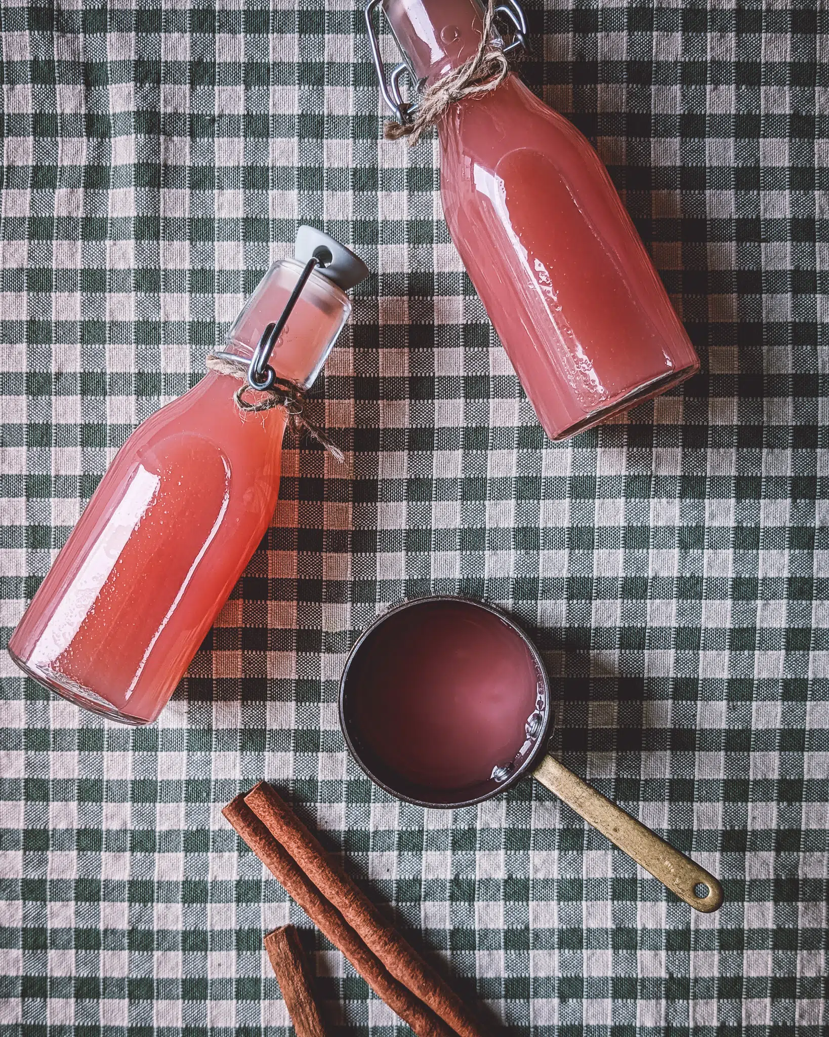 Two bottles of pink apple juice, a small pan with juice and a cinnamon twig on a green gingham tablecloth.