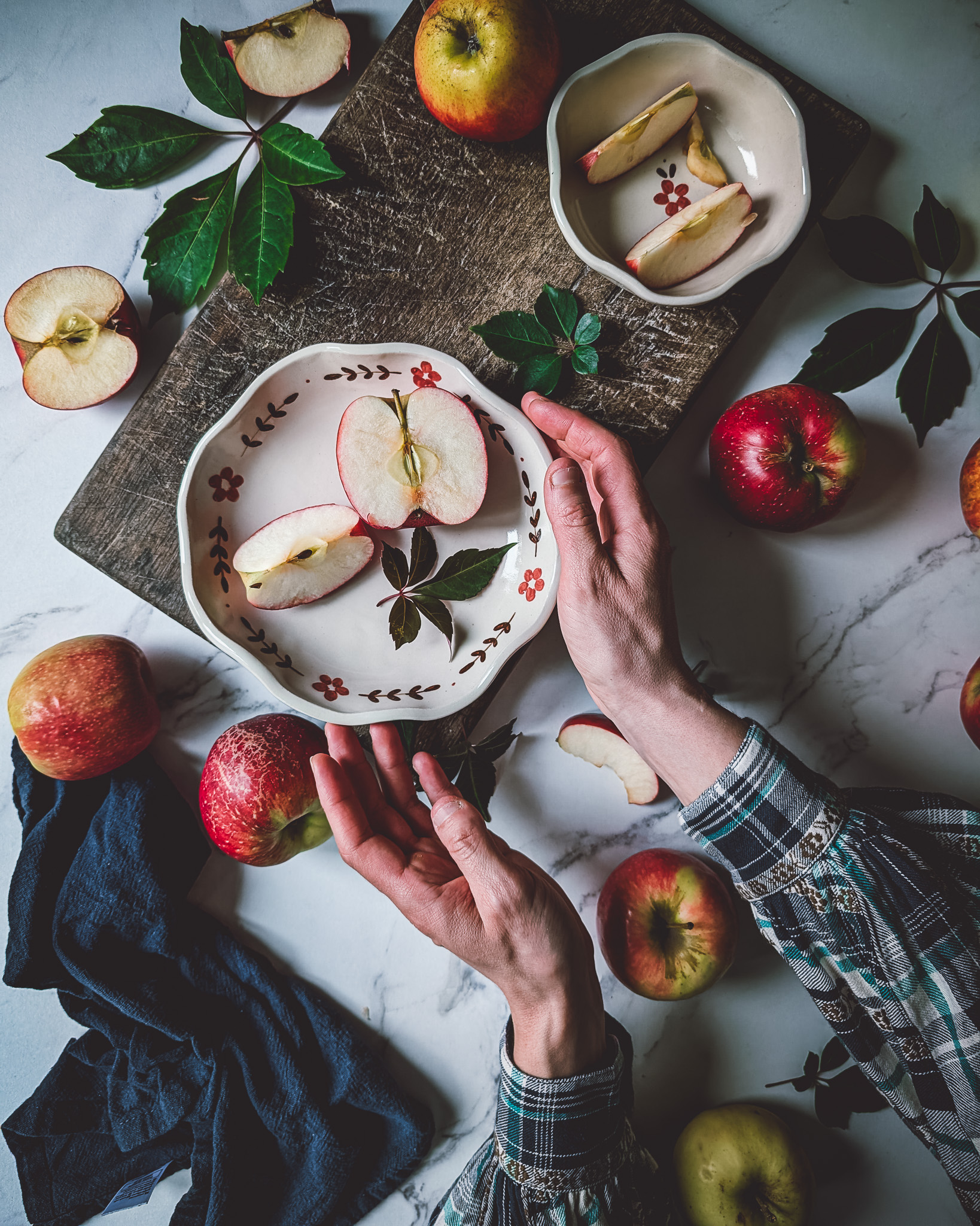Apples and chopped apples scattered on a marble table with cutlery, pretty dishes and a blue napkin. The hands of a person are lifting a place with an apple.