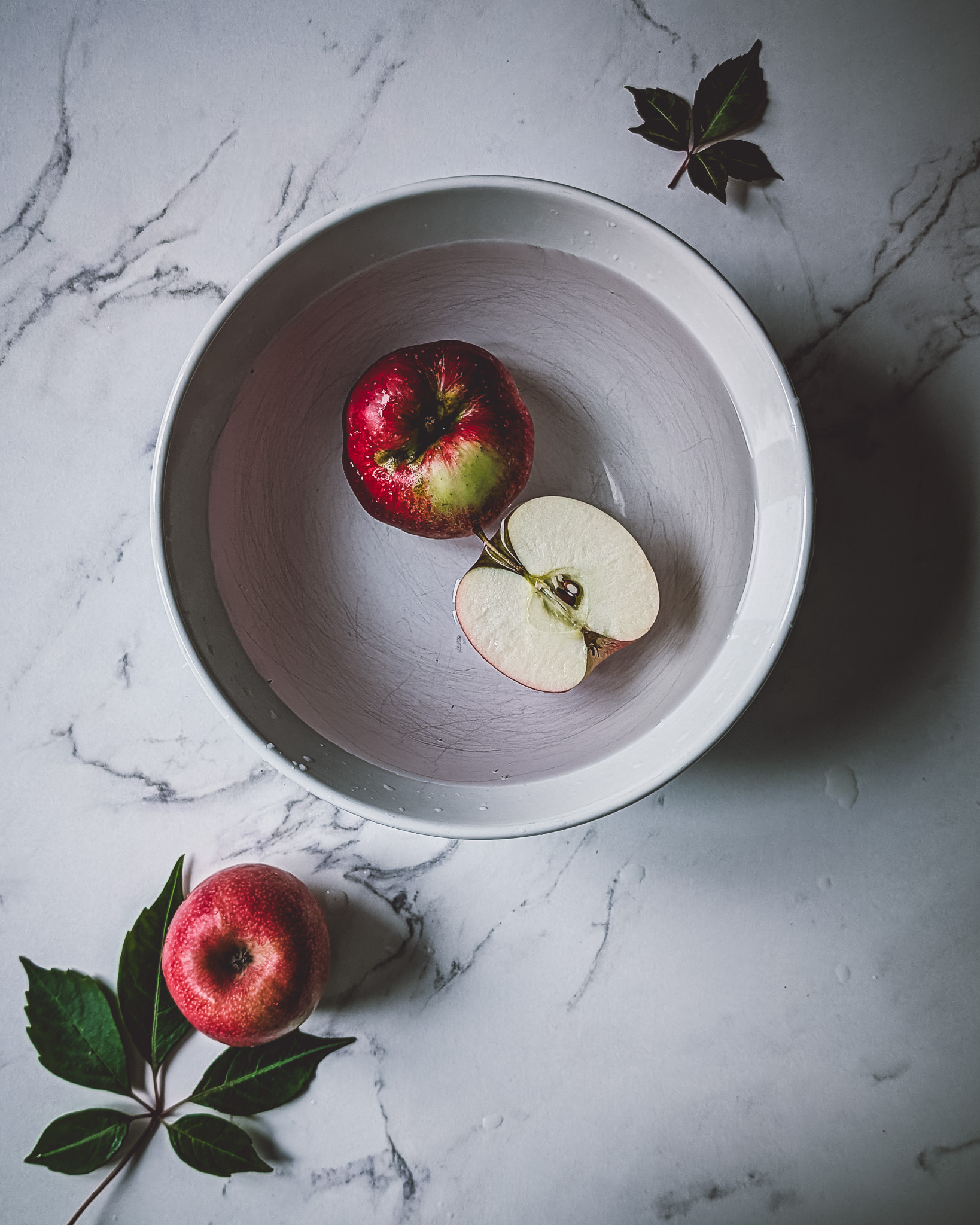 Two apples in a bowl full of water. On the table also a whole apple and some tree leaves.