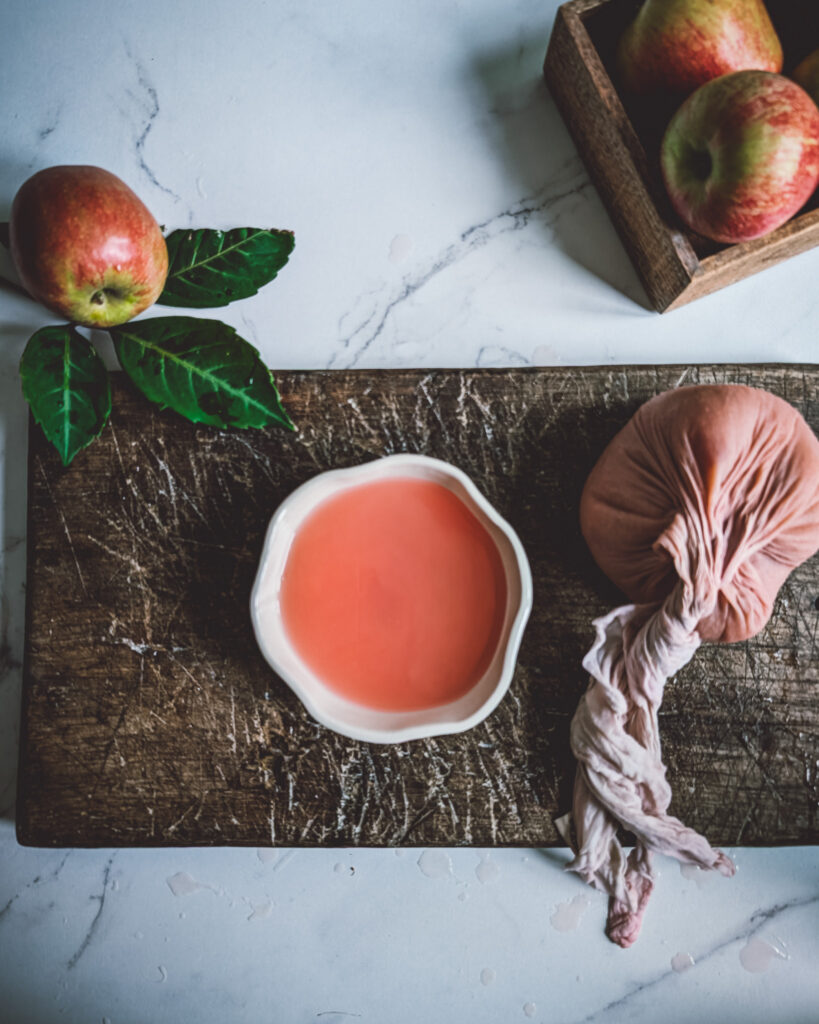A bowl full of apple juice and a muslim cloth full of freshly squeezed apple puree on a wooden board