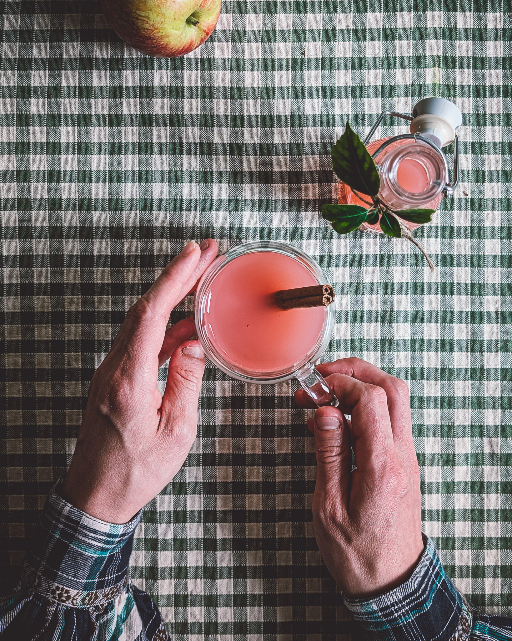 Hands of a person drinking from a transparent mug filled with apple juice. On the table also a small bottle of apple juice and a whole apple