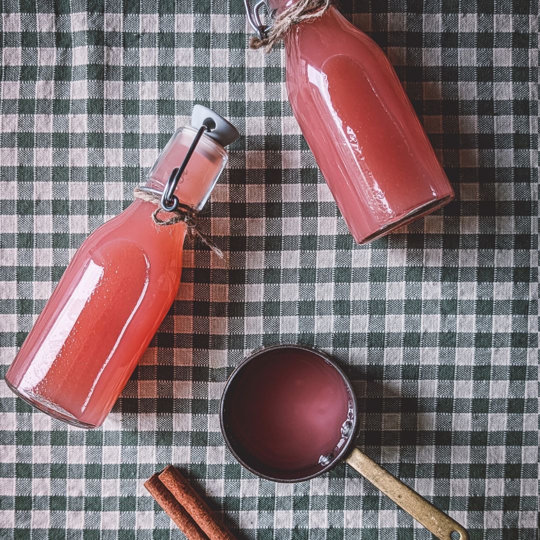 Two bottles of pink homemade apple juice, a small pan with juice and a cinnamon twig on a green gingham tablecloth.